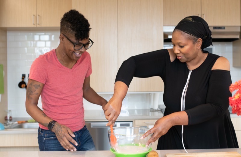 Two disabled Black people baking together in a kitchen. The woman on the right stirs a mixing bowl while the non-binary person on the left watches, laughing.
