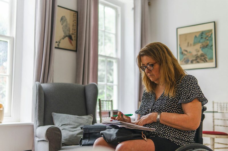 A woman in a wheelchair sits by a bright window at home, working through a sheaf of papers with a green pen.