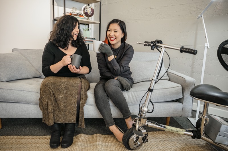 A Latinx disabled woman and an Asian disabled genderfluid person sit on a couch talking and laughing, both holding coffee mugs. A lightweight mobility scooter rests beside them.