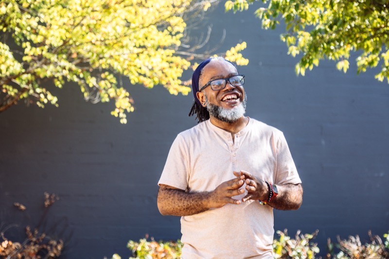 A Deaf Black man laughs in the middle of a conversation in sign language, standing outdoors in front of sunlit leaves.