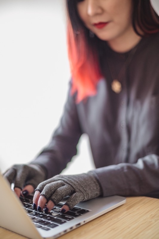 A disabled Asian genderfluid person types on a laptop wearing compression gloves. Their hands and the keyboard fill the frame.