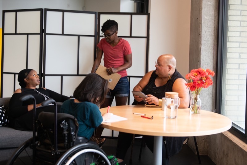 Four disabled people of colour meet around a table. One stands holding a large envelope, another writes in a notebook, and a third looks up from a tablet.