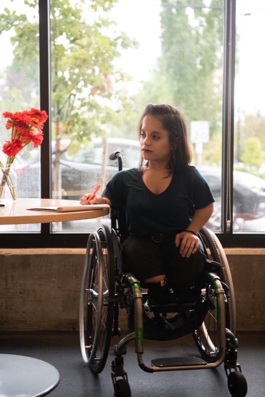 A South Asian disabled person in her wheelchair looks thoughtfully to one side while taking notes by hand, backlit by a window.