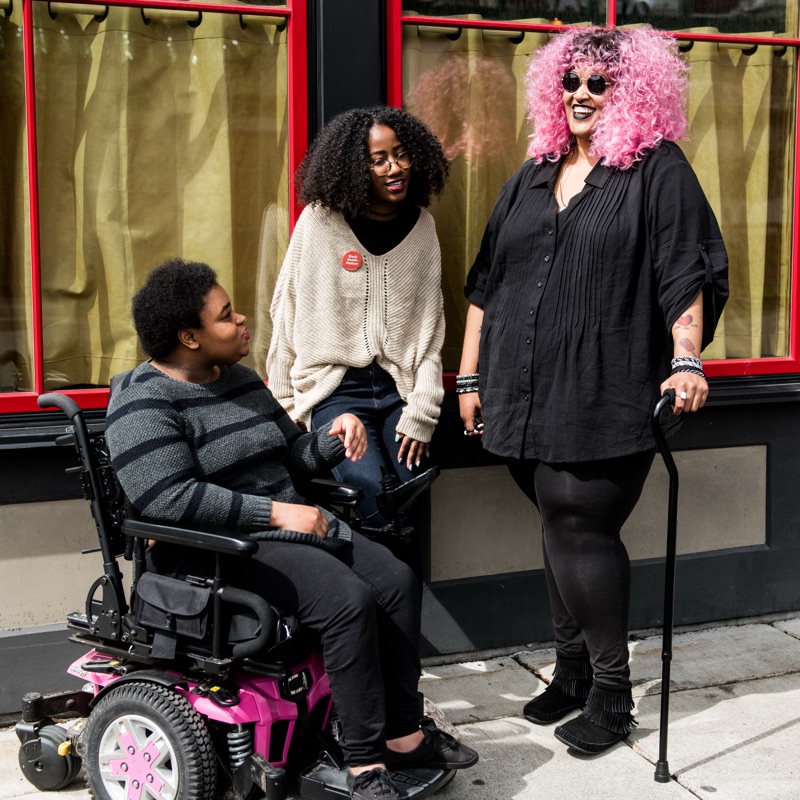 Three colleagues laughing together outside a building: one in a power wheelchair, one leaning against the window, one standing with a cane.